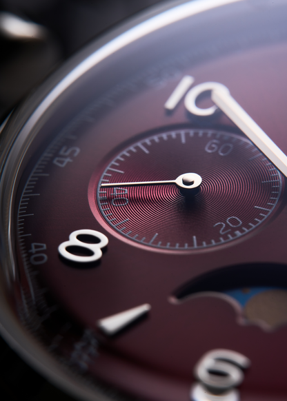Close-up of a wristwatch dial with a dark burgundy face, silver-tone numerals, and a smaller matching subdial.