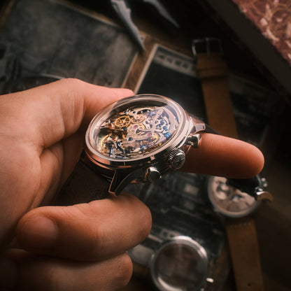 A close-up of a hand holding a skeleton wristwatch with a transparent case showing intricate gold and silver mechanical gears, with a dark leather strap.