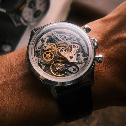 A close-up shot of a skeleton watch with a black strap worn on a wrist. The watch has a clear crystal case showing intricate gold and silver mechanical gears and a black leather strap.