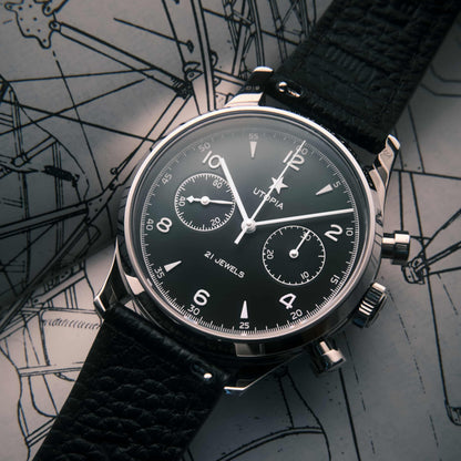 A close-up of a Utopia chronograph watch with a black dial, silver-tone case, and textured black leather strap, resting diagonally on a technical blueprint background.