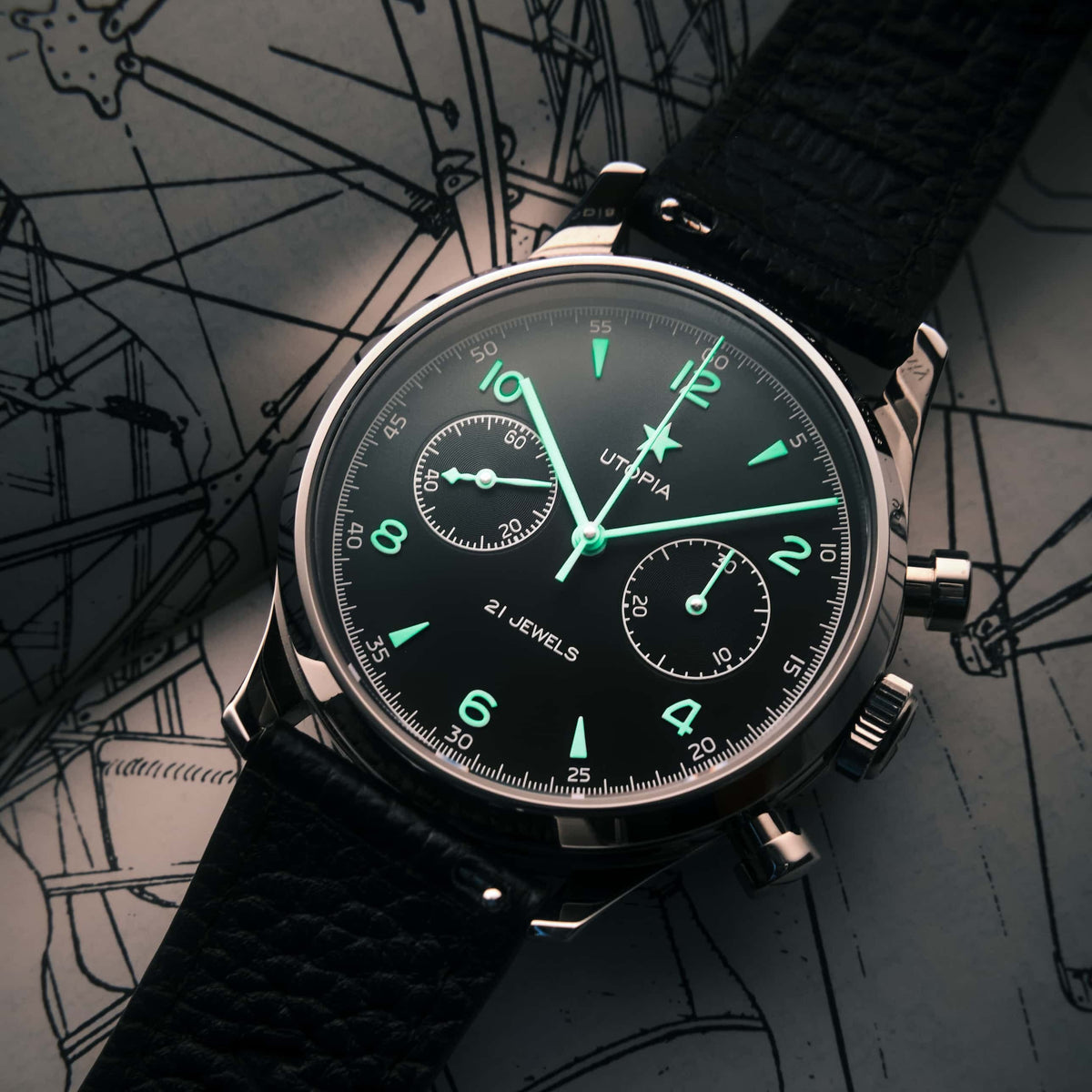 A close-up of a Utopia chronograph watch with a black dial, luminous green hands and markers, and a black textured leather strap, resting on a dark background with technical blueprint drawings.