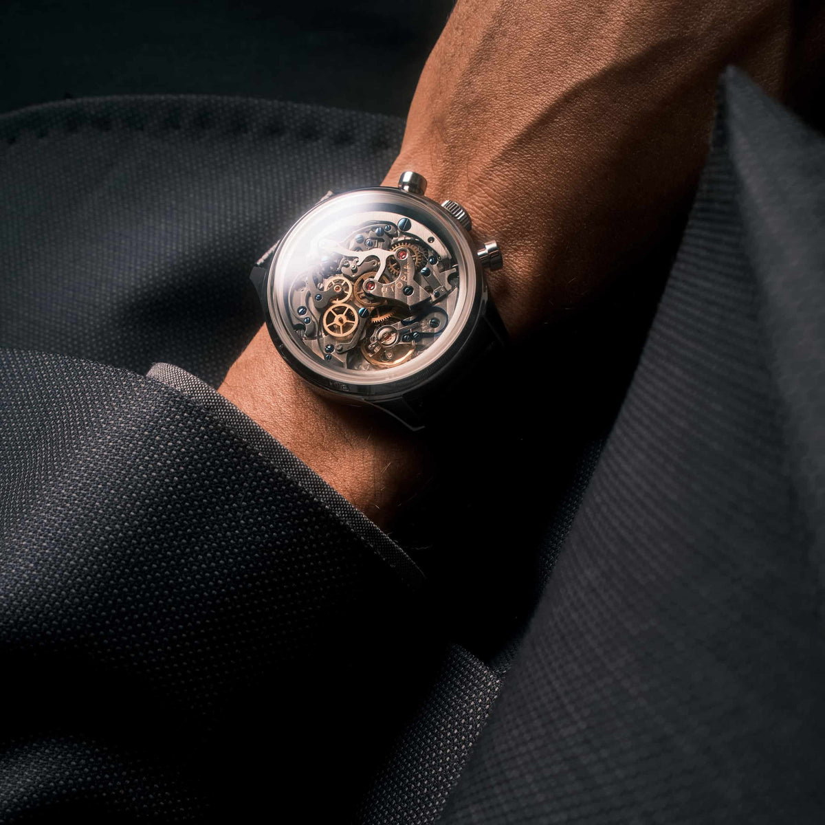 A close-up of a skeleton wristwatch with a silver-toned case and visible mechanical movement, worn on a person's wrist with dark clothing in the background.