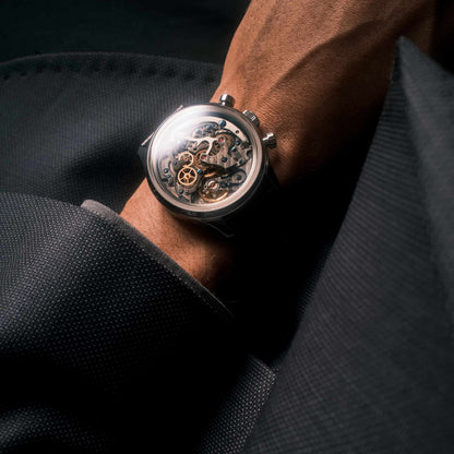 A close-up of a skeleton wristwatch with a silver-toned case and visible mechanical movement, worn on a person's wrist with dark clothing in the background.