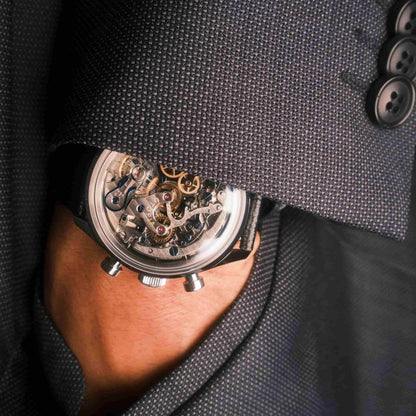 A close-up of a skeleton wristwatch with a black strap and silver-toned case, worn on a person's wrist and partially obscured by a dark textured suit jacket.
